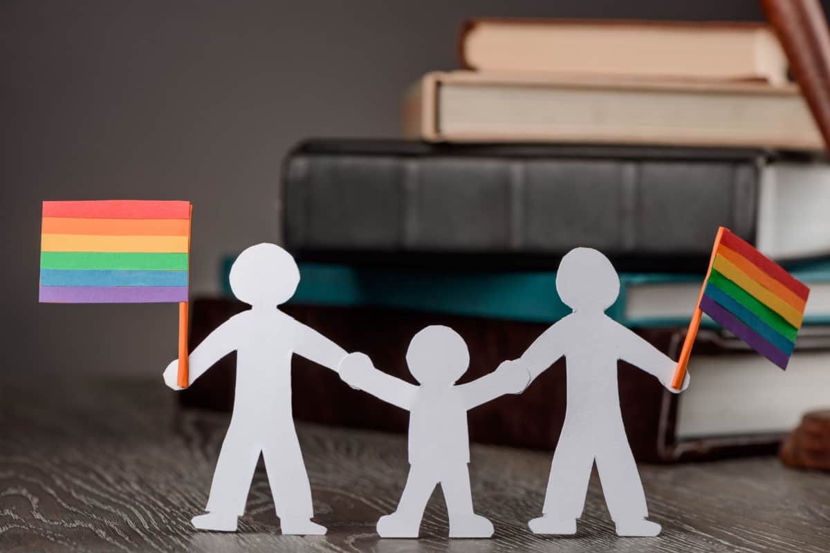 Paper cutouts of a family holding hands and holding small rainbow pride flags against a blurred background of law books.
