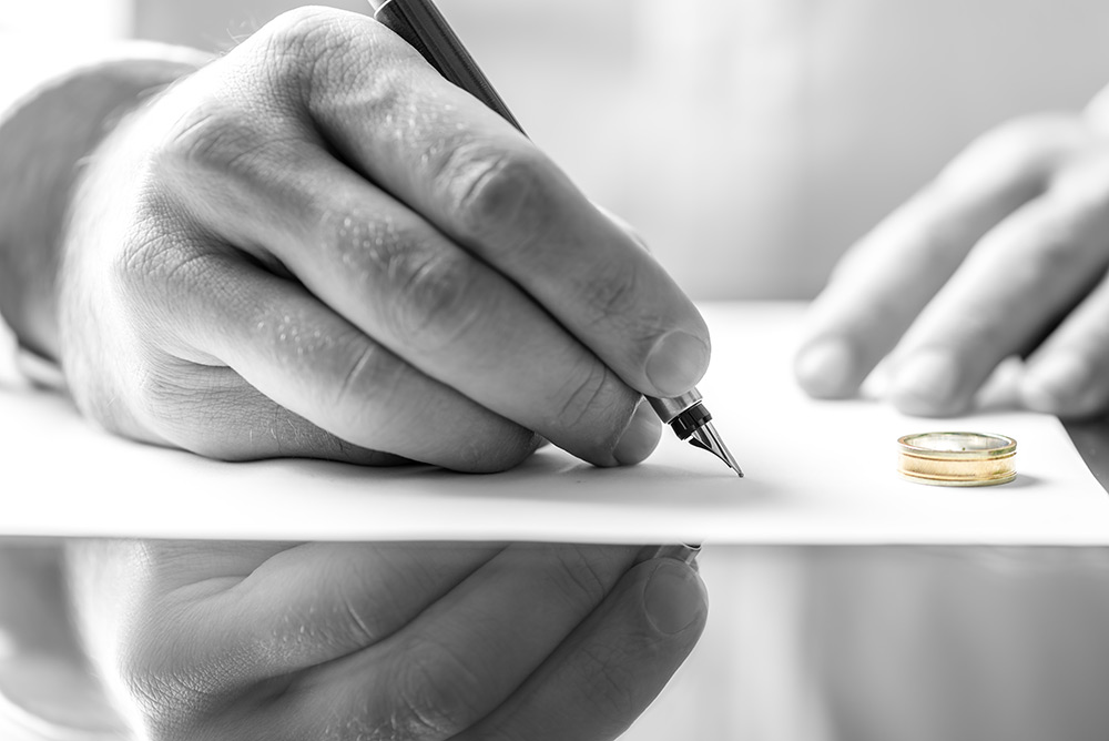 Close-up of a person signing legal documents with a fountain pen next to a gold wedding ring on a desk.